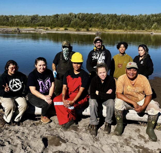 A diverse group of nine people dressed in work clothes pose for a photo on the river's shoreline.